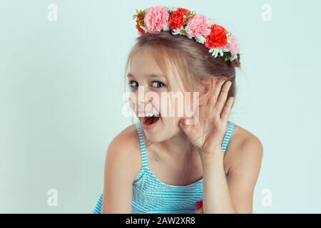 Joyeux enfant fille main à oreille écoutes essayer d'entendre à un chichat. Gros portrait du modèle caucasien pour enfant avec bandeau floral isolé sur le blanc Banque D'Images