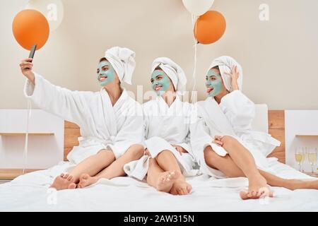 Vue avant du groupe souriant de filles avec masques cosmétiques, serviettes sur la tête et peignoirs s'amuser à la maison. Trois jolies filles amis prenant selfie dans la chambre avec des ballons, champagne à proximité. Banque D'Images