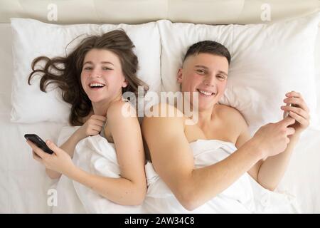 Vue de dessus d'un couple souriant allongé dans le lit avec un smartphone. Banque D'Images