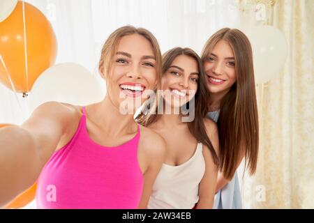 Gros plan sur un groupe souriant de jeunes jolies filles dans des robes prenant le selfie. Trois amis émotionnels de sexe féminin qui s'amusent à la fête à domicile, célébrant. Concept d'amitié, d'humeur festive. Banque D'Images