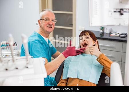 Belle jeune femme au bureau du dentiste satisfait après un traitement dentaire réussi, assis sur la chaise et montrant le pouce avec le haut Banque D'Images