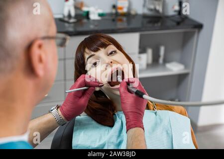 Jeune femme caucasienne, assise en chaise de dentiste, regardant la caméra à bouche ouverte tout en vérifiant les dents par le médecin. Banque D'Images