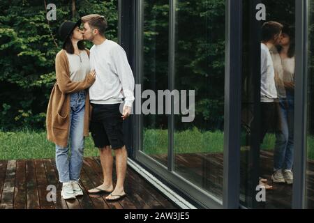 Élégant couple hipster embrassant et embrassant sur la terrasse en bois, se détendre dans la cabine dans les montagnes. Une famille heureuse en costume moderne embrassant sur le dos Banque D'Images