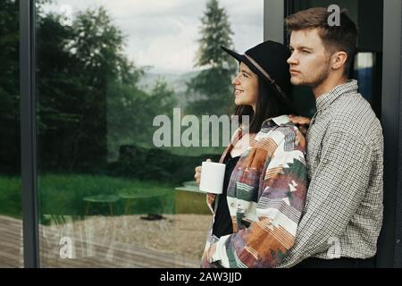 Élégant couple hipster avec café du matin reposant sur l'arrière-plan de la cabine moderne et de grandes fenêtres dans les montagnes. Bonne famille à profiter du matin à Banque D'Images