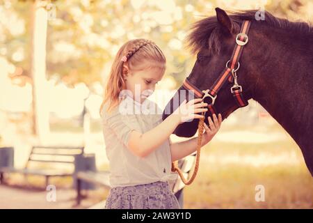 Mon ami charmant. Gros plan portrait mignon petite fille tenant câlin son cheval poney isolé dehors parc vert arrière-plan au coucher du soleil Banque D'Images