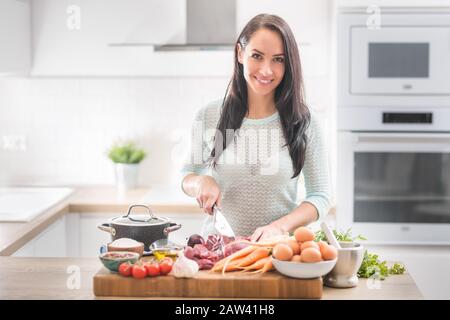 La jeune femme gaie est heureuse de préparer le déjeuner dans sa cuisine. Elle prépare des aliments à partir de viande et de légumes et d'autres ingrédients sains. Banque D'Images