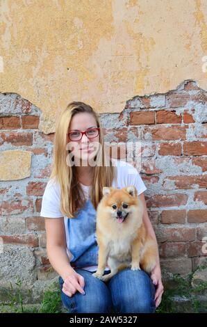 Jeune fille d'à côté mignon portant des lunettes rouges, un Jean et un t-shirt, posant avec son animal de compagnie devant le mur de briques grunge Banque D'Images