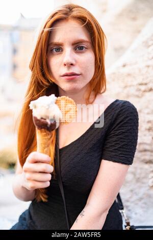 Jolie jeune fille au gingembre calme, vêque d'une robe noire, assise dans la rue de la ville avec un cône de crème glacée à la main et regardant l'appareil photo Banque D'Images