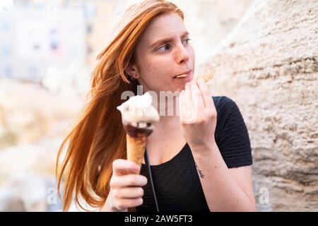 Jolie fille millénaire avec de longs cheveux rouges, un délicieux cône de glace avec biscuit et regardant loin tout en étant assise dans la rue de la ville en été Banque D'Images
