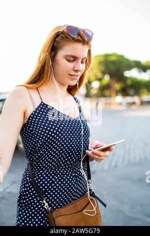 Une jeune dame heureuse avec des cheveux rouges souriant et utilisant un smartphone tout en se tenant dans la rue de la ville et en écoutant de la musique le jour ensoleillé Banque D'Images