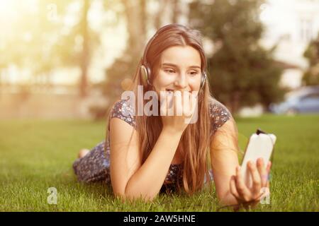 Regarder un film à l'extérieur. Happy Woman adolescente femme regardant le téléphone cellulaire piquer avec anxiété les ongles en profitant des écouteurs de jeu sur la tête souriant Banque D'Images