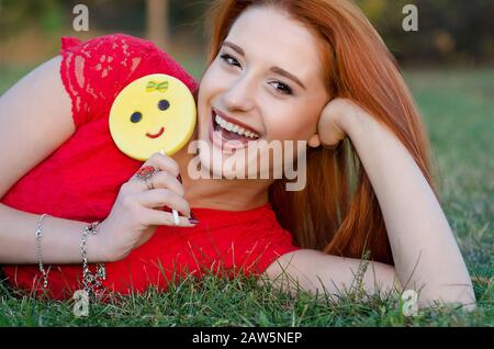 Charmante fille à tête rouge avec des bonbons à sucre repose sur l'herbe verte et rie joyeusement Banque D'Images