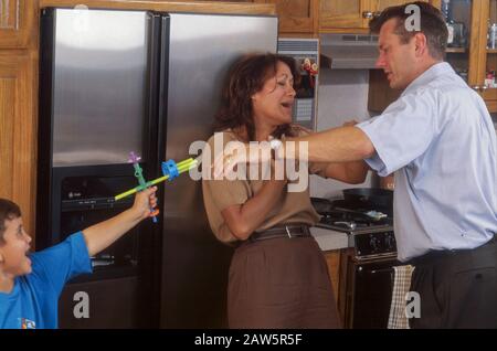 La photo posée de petit garçon pointe son fusil de jouet à ses parents pendant qu'ils soutiennent, illustrant le comportement appris. Banque D'Images