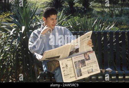 Un homme hispanique de 18 ans lit le journal de langue espagnole tout en étant assis sur un banc de parc public à Austin, Texas. Banque D'Images