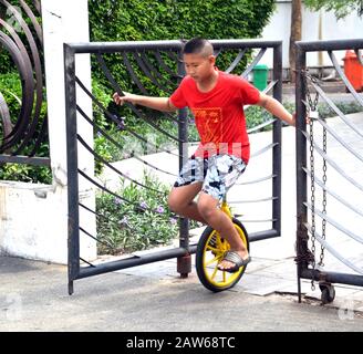 Un jeune garçon asiatique, âgé de 12 à 13 ans, pratique à cheval sur un monocycle, se tenant d'abord à une porte métallique pour soutenir se stabiliser, à Bangkok, en Thaïlande, en Asie du Sud-est. Banque D'Images