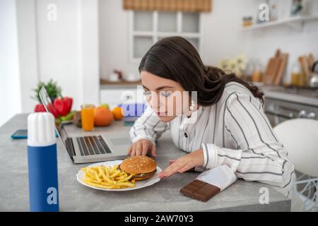 Femme aux cheveux sombres dans un chemisier à rayures mangeant des hamburgers et des chips de pommes de terre Banque D'Images