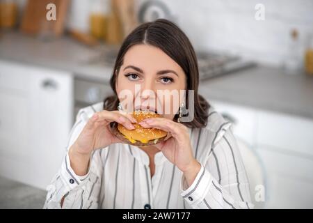 Femme aux cheveux sombres dans un chemisier à rayures mangeant un gros hamburger Banque D'Images