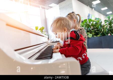 Adorable petit garçon caucasien mignon ayant plaisir à jouer un grand piano blanc dans un centre commercial intérieur. Un petit enfant joyeux et drôle aime chanter et faire du bruit Banque D'Images