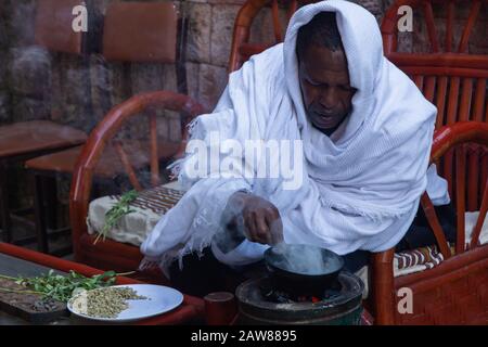 Lalibela, Ethiopie - Nov 2018: Homme vêtu de vêtements éthiopiens blancs traditionnels torréfaction des grains de café avant de préparer un café. Banque D'Images