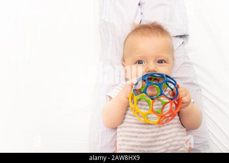 Un petit mignon petit bébé joue avec une boule en plastique colorée posée sur le dos et tenir le jouet près de la bouche Banque D'Images