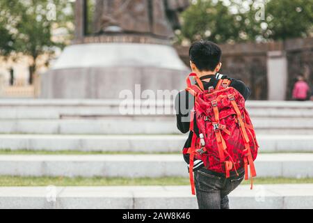 Un jeune photographe asiatique masculin avec un sac à dos brillant sur ses épaules photographie le monument. Vue arrière Banque D'Images