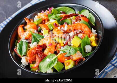 salade d'épinards aux crevettes avocat avec tomates séchées au soleil, morceaux de bacon frits, fromage cotija émietté, oignon rouge dans un bol noir sur une table en béton, clos Banque D'Images