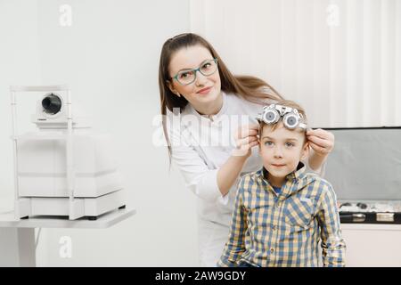 Jeune fille médecin tenant des lunettes spéciales de patient pour les tests oculaires et la sélection des lentilles. Petit beau garçon blond avec de gros yeux bleus long des paupières Banque D'Images