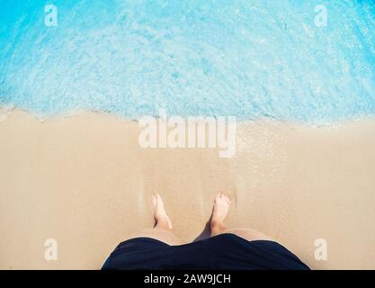 Les pieds de pied d'homme dans le sable doré sont lavés par des vagues de mer bleue, vue de dessus. Concept de freelance de voyage indépendant. Banque D'Images