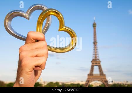 Main tenant paire de coeurs d'or et d'argent à l'extérieur devant une vue ensoleillée de la Tour Eiffel à Paris, France Banque D'Images