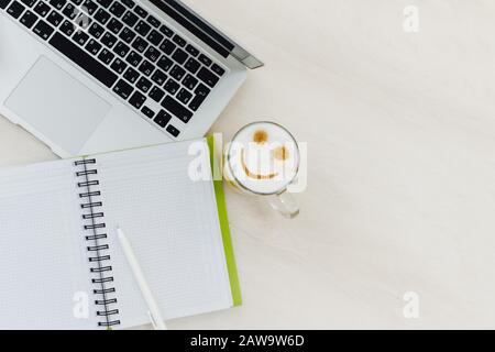 Un café du matin revigorant avec un motif émoticône sur une table en bois clair à côté d'un ordinateur portable et un cahier vierge. Modèle de blog. Copyspace. Banque D'Images