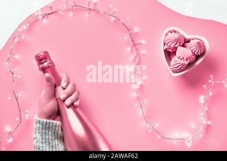 Vue sur le dessus de la Saint-Valentin sur fond rose. Guirlande lumineuse, mains de femme montrant le signe du cœur. Bouteille de champagne rose métallisé. Disposition plate monochrome tendance dans vi Banque D'Images