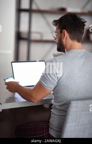 jeune homme adulte avec barbe portant des lunettes, regardant son téléphone portable devant l'ordinateur portable sur la table. technologie, style de vie, freelance, médias sociaux conc Banque D'Images