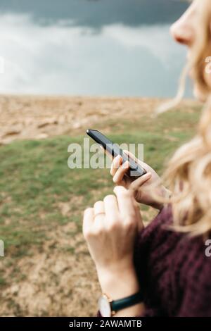 Gros plan photo des mains féminines tenant le téléphone mobile et prenant des photos de lac sur un jour nuageux. Elle envoie des SMS sur son smartphone et prend son selfie. Banque D'Images