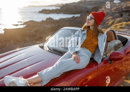 Jeune femme élégante qui profite de paysages magnifiques, allongé sur le capot de la voiture tout en voyageant sur la côte rocheuse au coucher du soleil. Style de vie sans souci et concept de voyage en voiture Banque D'Images