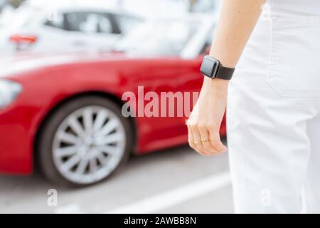 Femme avec montre intelligente au parking avec véhicule rouge sur l'arrière-plan, vue rapprochée sur montre avec écran noir. Contrôle de la voiture avec un concept de montre intelligente Banque D'Images