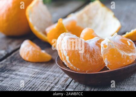 Des tranches de mandarine jaune mûre se trouvent dans un bol près de la peau de mandarine sur une table en bois. Agrumes, mandarines ou oranges. Gros plan. Banque D'Images