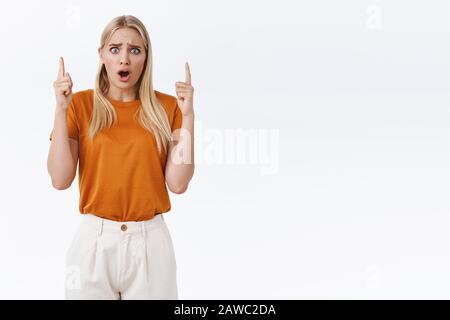 Une jeune fille blonde soyeuse et affligée, en t-shirt orange élégant, frognant, inquiète et déçue, en faisant pointer les doigts vers le haut pour réagir Banque D'Images