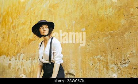 Une fille dans un chapeau se tient par le mur et regarde autour. Grand vieux mur jaune. Banque D'Images