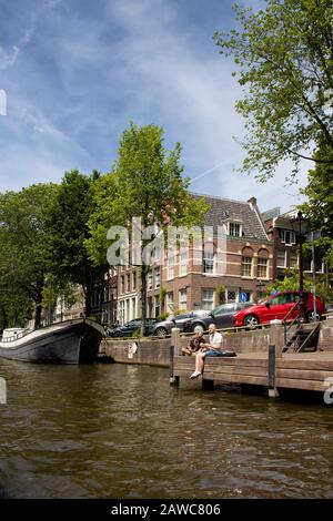 Vue sur deux hommes penchant par canal, arbres, voitures garées et un bateau, bâtiments historiques et traditionnels montrant le style architectural néerlandais à Amsterdam Banque D'Images