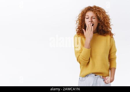 Cette jeune femme moderne aux cheveux bouclés se tient debout au bureau, prépare du café le matin, porte un pull jaune, un yawn et couvre la bouche ouverte Banque D'Images