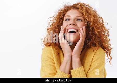 Une femme rêveuse et enchantée à tête rouge à poil vif dans un pull jaune, touchant sa peau pure et propre et souriante, riant avec joie, regardant vers le haut Banque D'Images