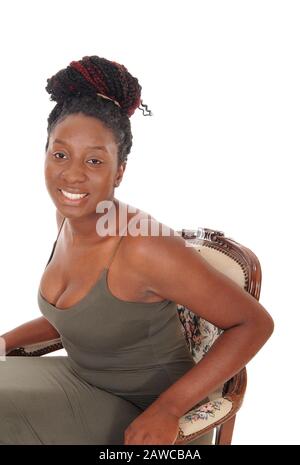 Une heureuse femme africaine souriante assise dans une robe grise dans un vieux fauteuil, regardant dans l'appareil photo, isolée pour un fond blanc Banque D'Images