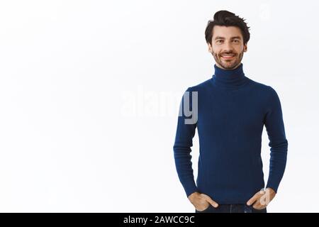 Taille-up portrait confiant homme barbu portant élégant pull bleu à col haut, tenir les mains dans les poches et sourire arrogant, chic debout Banque D'Images