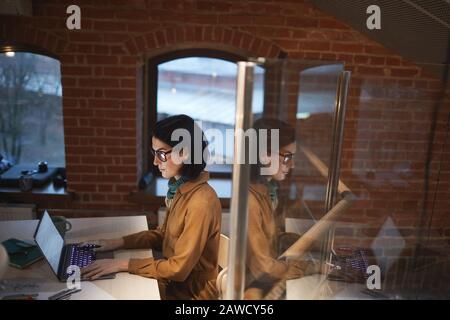 Jeune femme d'affaires dans des lunettes assis à la table devant l'ordinateur portable et travaillant sur lui au bureau moderne Banque D'Images