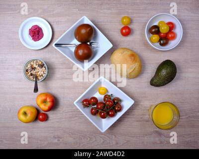 Bretzel, gâteau sucré au thé, marmelade, avocat, tomates, grenade et verre de jus d'orange, disposés symétriquement sur une table en bois sur blanc et Banque D'Images