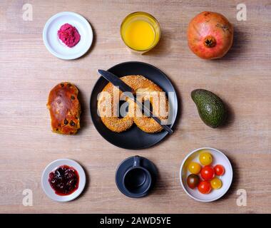 Bretzel, gâteau sucré au thé, marmelade, avocat, tomates, grenade et verre de jus d'orange, disposés symétriquement sur une table en bois sur blanc et Banque D'Images