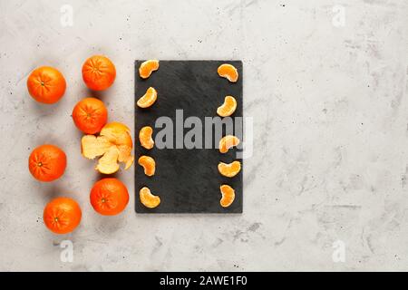 Des tranches de mandarine pelées, vives et juteuses, se trouvent sur une ardoise grise, sur un fond de béton gris, des fruits orange entiers se trouvent à proximité au hasard. Banque D'Images