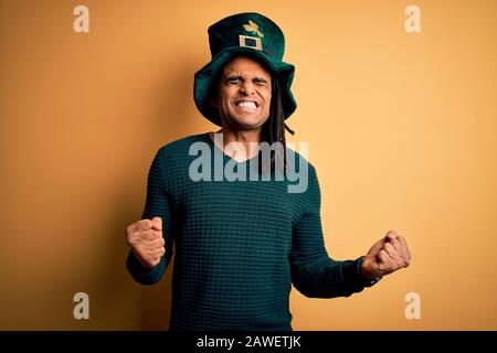 Jeune homme africain américain portant un chapeau vert avec un trèfle célébrant saint patricks jour excité pour le succès avec les bras levés et les yeux fermés célébrati Banque D'Images