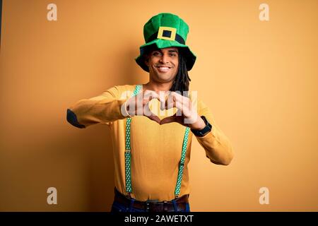 Jeune homme africain américain portant un chapeau vert célébrant la Saint patricks jour souriant en amour faisant la forme de symbole de coeur avec les mains. Concept romantique. Banque D'Images