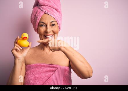 Femme d'âge moyen portant une serviette de bain rose des soins du corps de beauté tenant le canard jaune en caoutchouc très heureux pointant avec la main et le doigt Banque D'Images